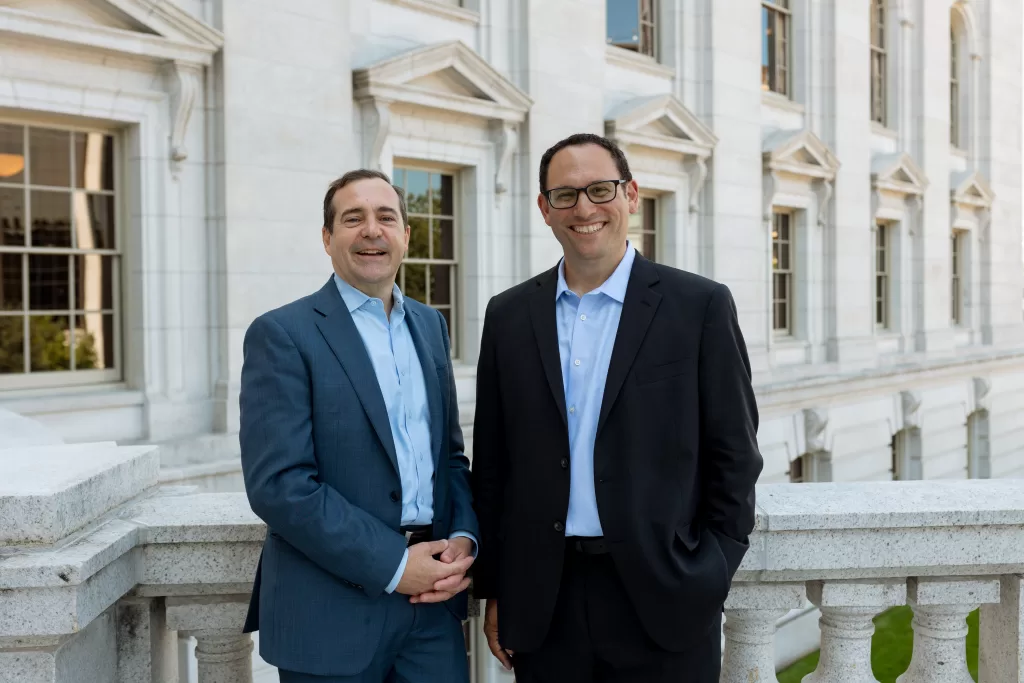 Doug Poland and Jeff Manzel standing in front of the Wisconsin Capitol building, both wearing suits with blue shirts.